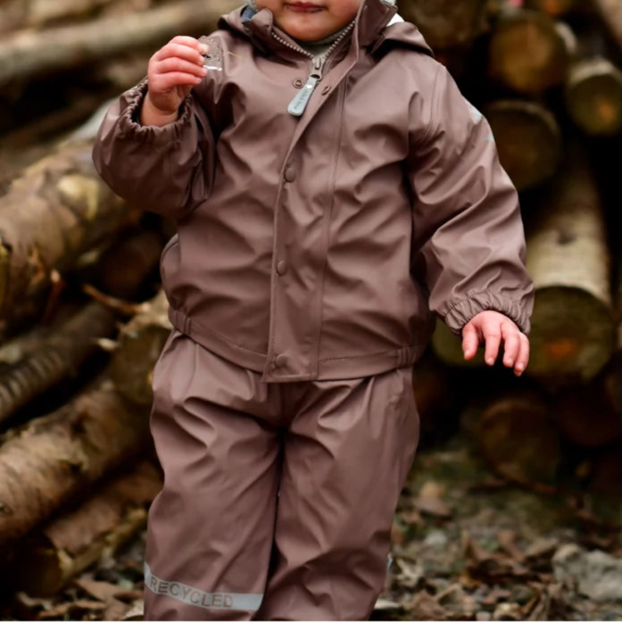 Child wearing a brown raincoat and pants standing in front of stacked logs.