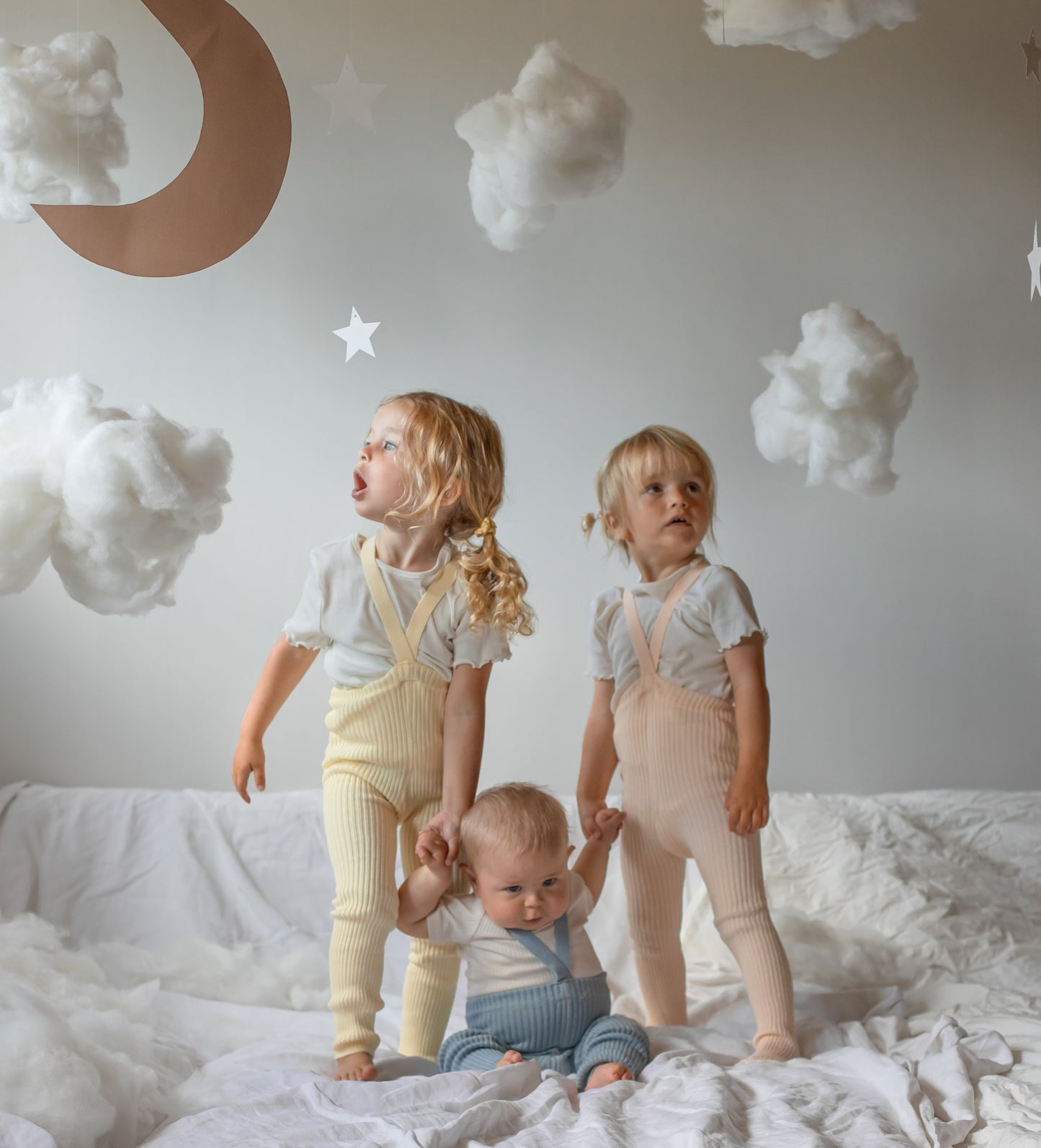 Three children standing and sitting on a rumpled white bed in a softly lit nursery, wearing pastel ribbed overalls in cream, pale yellow, and light blue. The room has white clouds, stars, and a crescent moon wall decor.